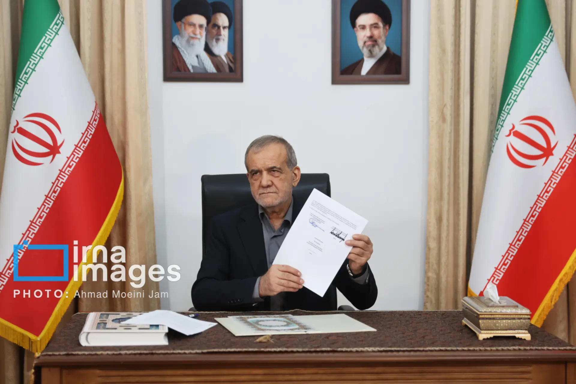 مسعود پزشکیان در حال گرفتن عکس یادگاری با سند تفاهم جمهوری اسلامی و امریکا زیر عکس رهبران جمهوری اسلامی