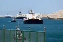 چندین کشتی عظیم تجاری و نفتکش در حال حرکت در آب‌های آبی کانال سوئز در کنار ساحل بیابانی.