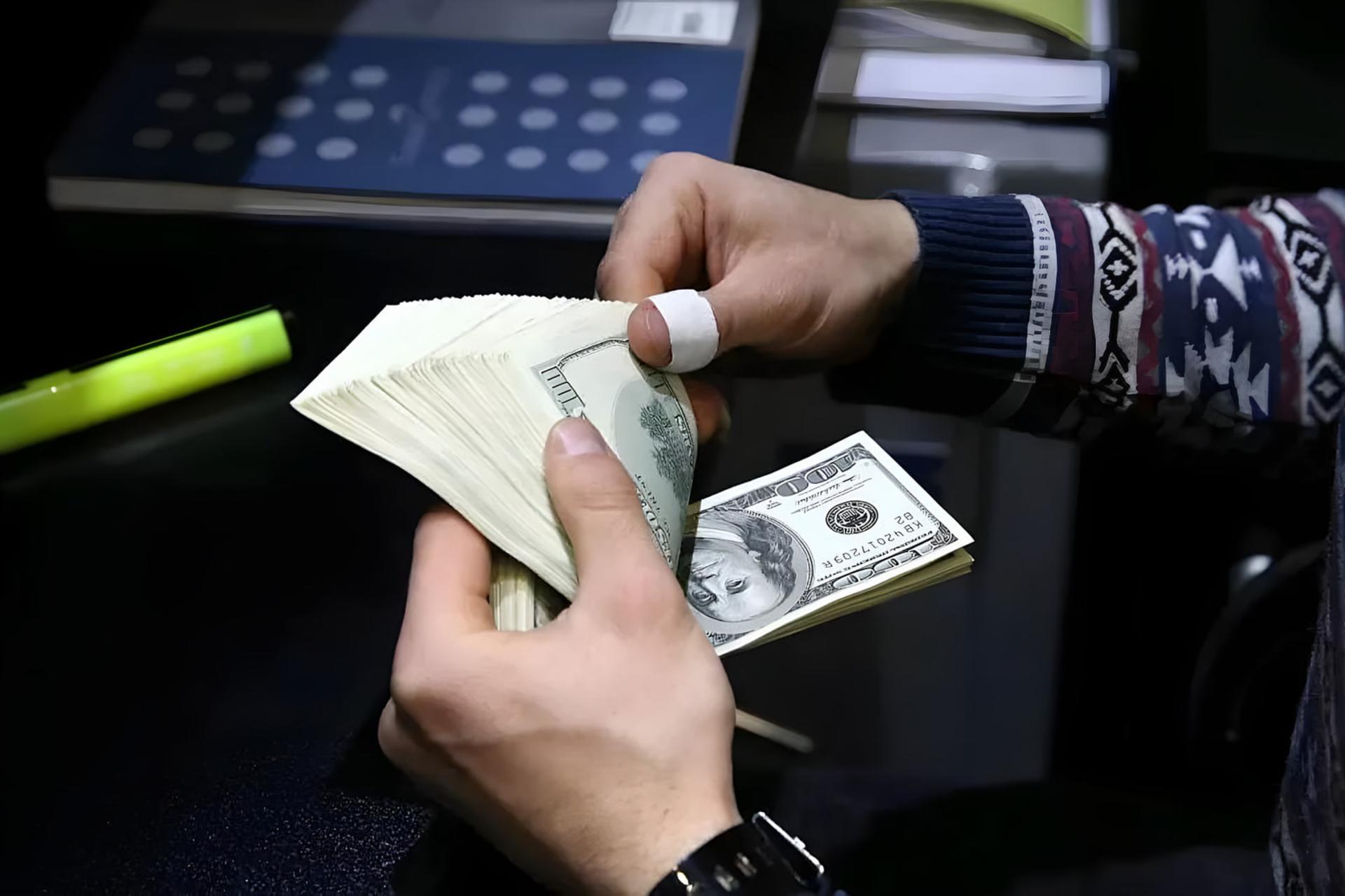 دست‌های فردی در حال شمارش دسته‌های اسکناس دلار (US Dollar) در محیطی تاریک