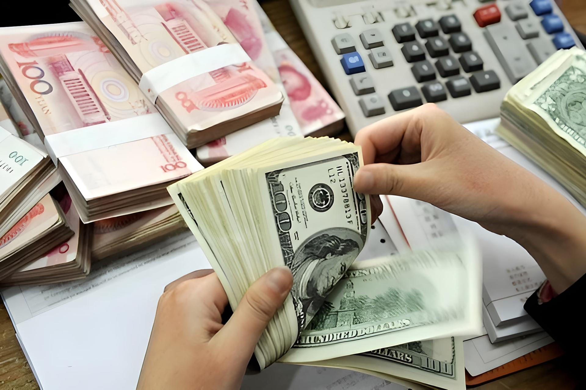 دست‌های فردی در حال شمارش اسکناس‌های دلار آمریکا در کنار بسته‌های پول یوآن