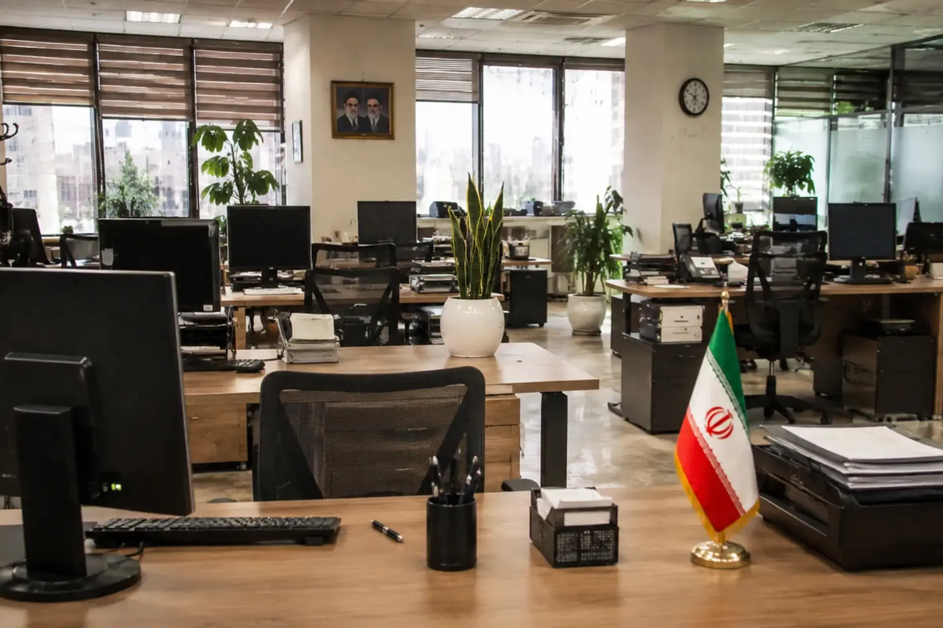 میزهای کار اداری با مانیتور و پرچم ایران در محیط دفتر
