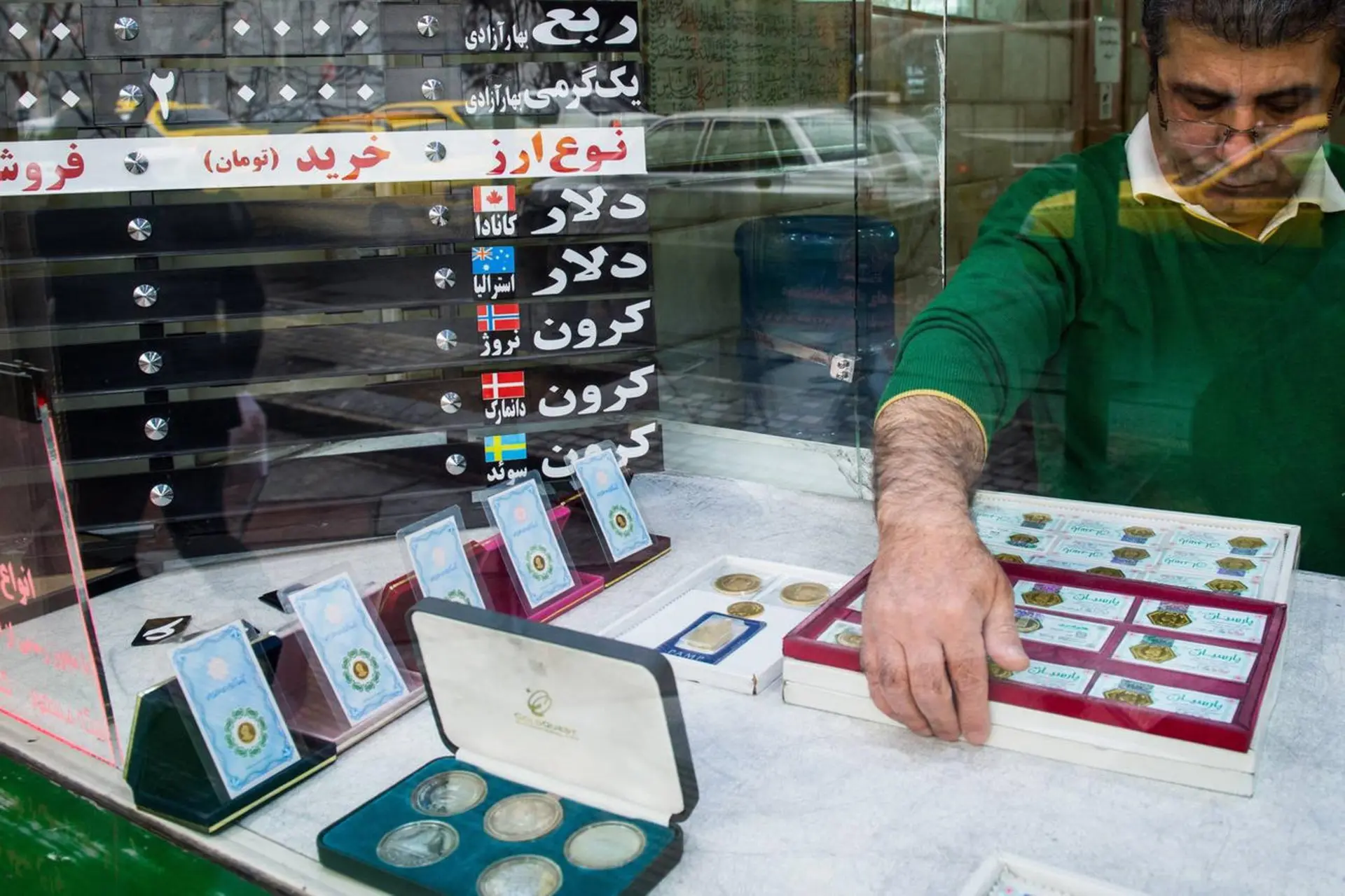 مردی در حال چیدمان سکه‌ها و اسکناس‌های قدیمی در ویترین یک مغازه تعویض ارز است.