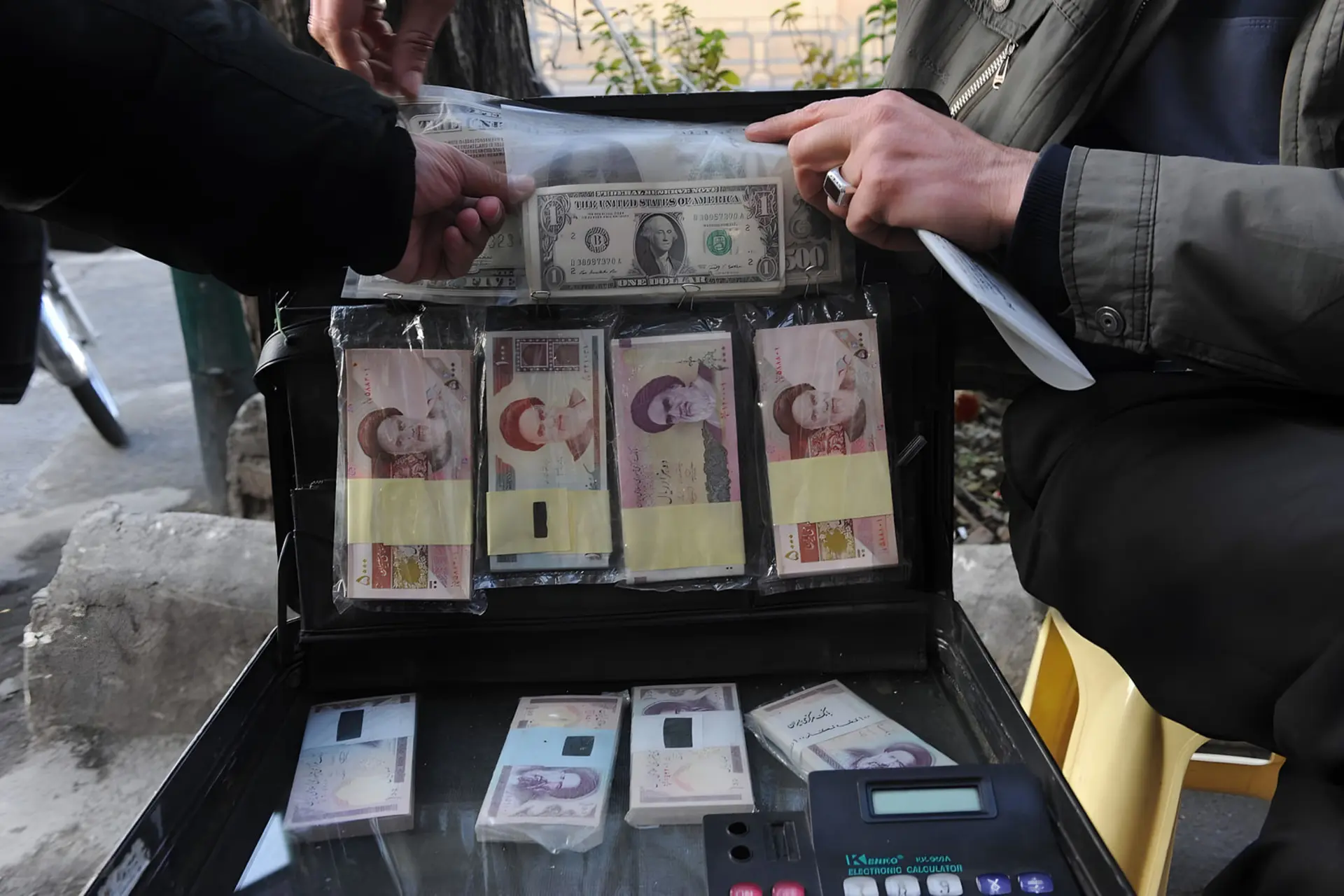 دست‌های افراد در حال جابه‌جایی بسته‌های پول نقد دلار (USD) و ریال ایران