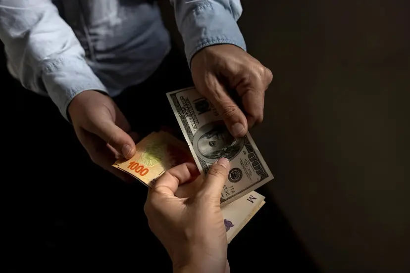 دست‌های دو نفر در حال رد و بدل کردن اسکناس‌های ۱۰۰ دلاری (USD) و پول محلی