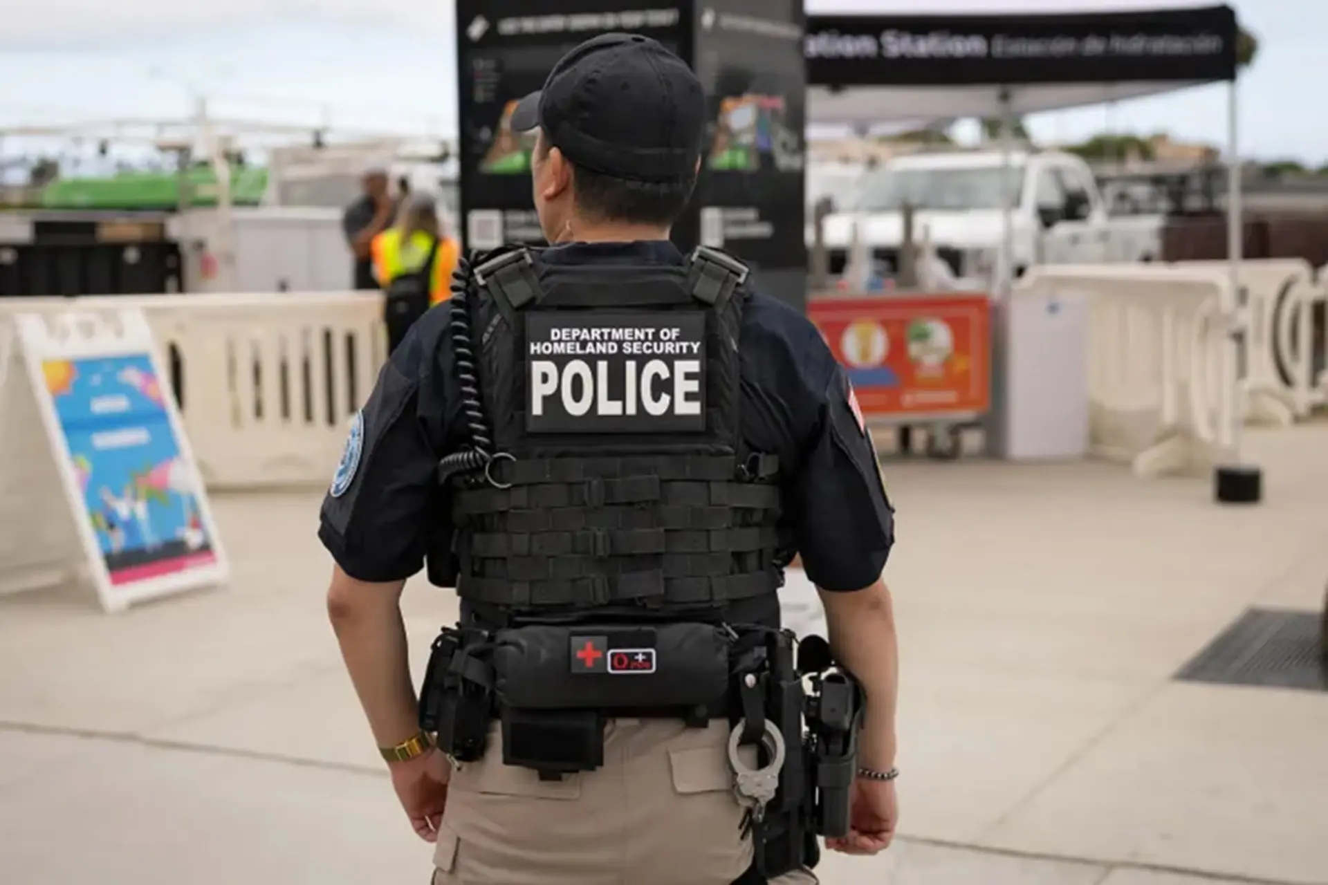 نمای پشت یک افسر پلیس با جلیقه تاکتیکی نوشته شده Department of Homeland Security Police در یک محیط باز.