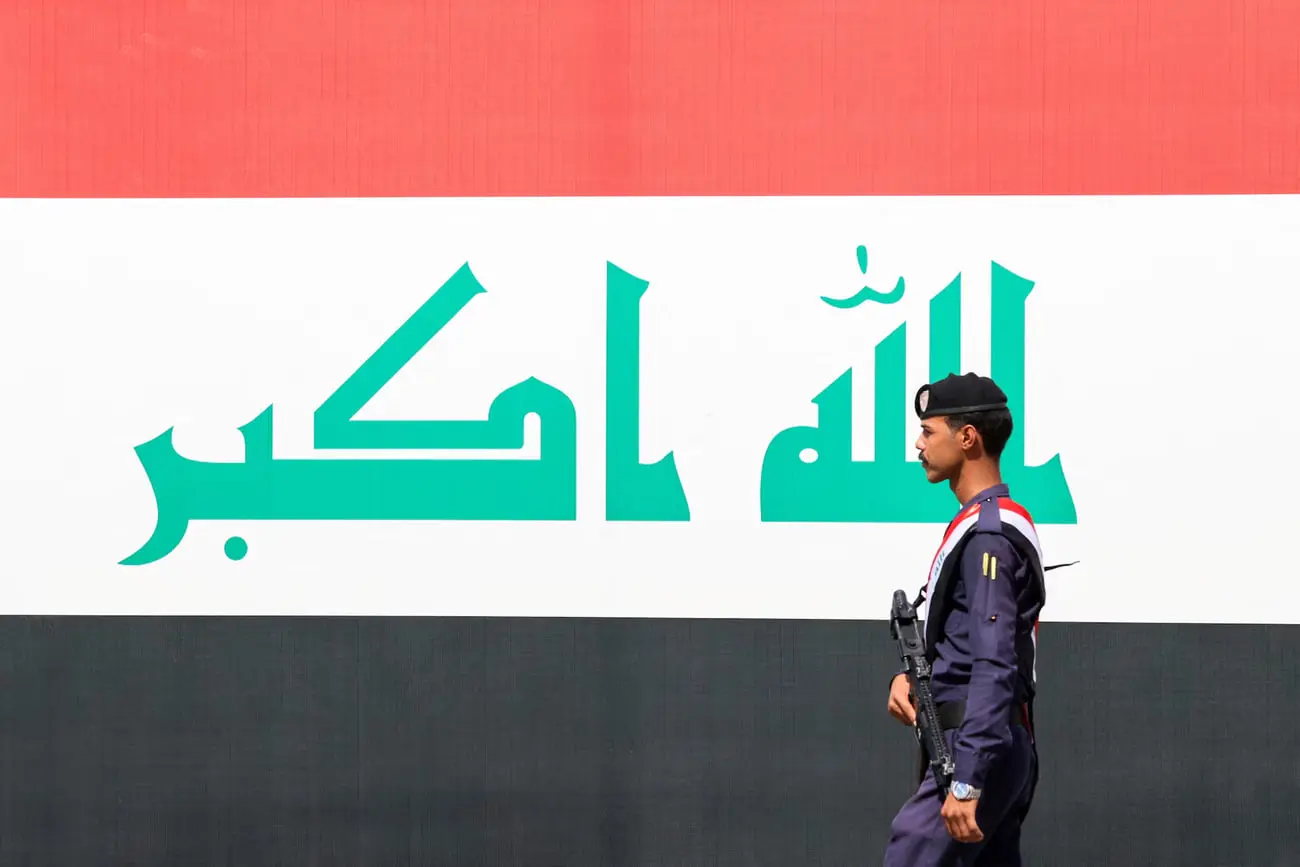پرچم عراق (Iraq flag) با نوشته‌های سبز در پس‌زمینه و سربازی با لباس نظامی