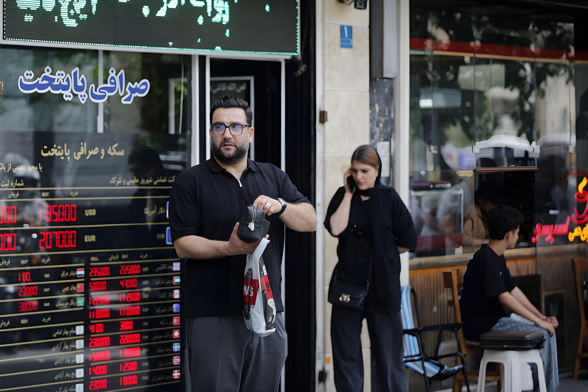 مردی با لباس مشکی در حال ایستادن مقابل ویترین یک صرافی با نمایشگر قیمت ارز
