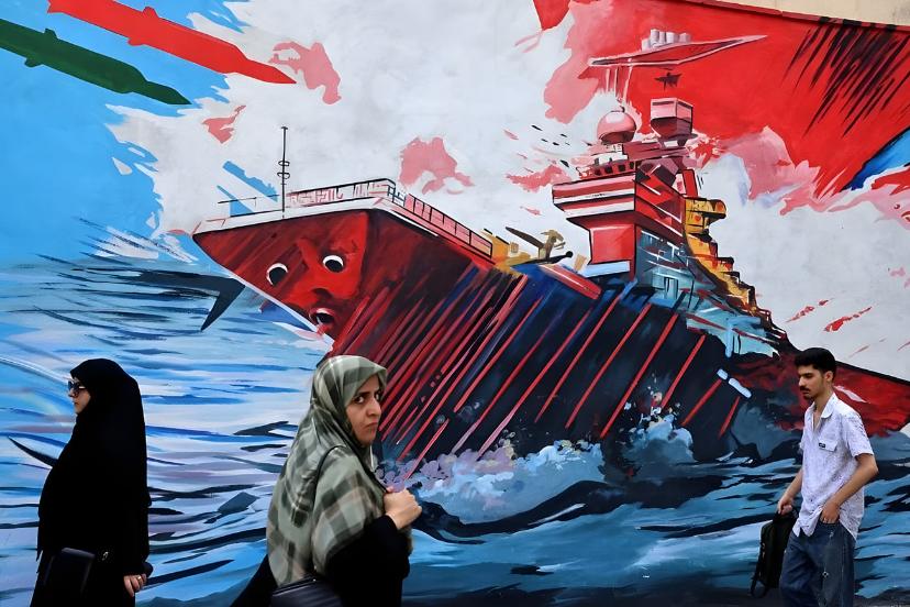 سه عابر در حال عبور از دیوار نگاره ای در تهران که محتوایش حمله به ناو امریکایی است