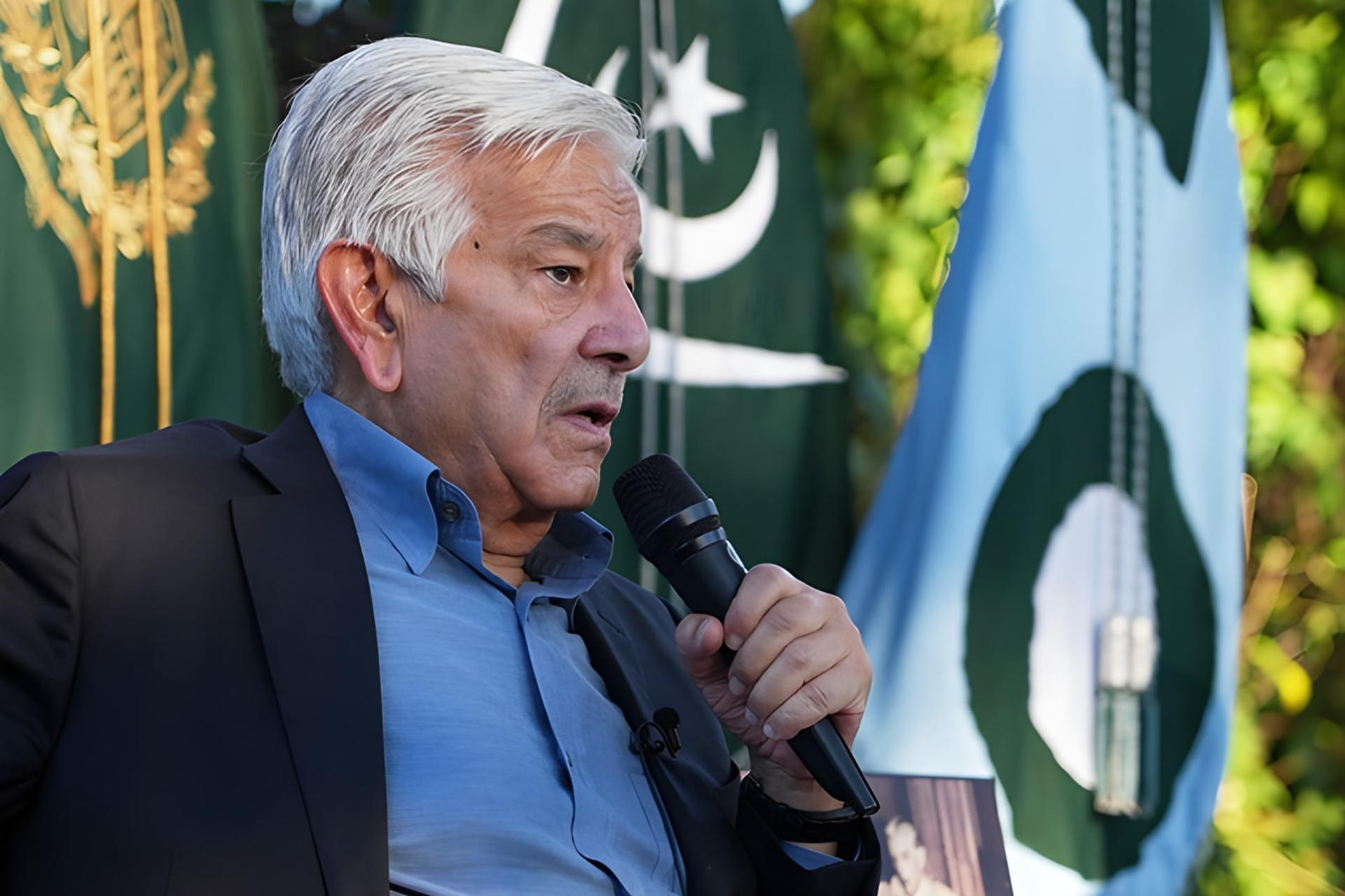 مردی با موهای سفید در حال سخنرانی با میکروفون در کنار پرچم پاکستان
