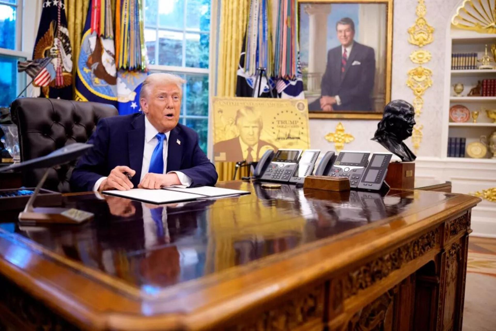 دونالد ترامپ پشت میز کار رسمی در یک کاخ سفید نشسته است. در پس زمینه عکس ریگان نیز دیده می شود.