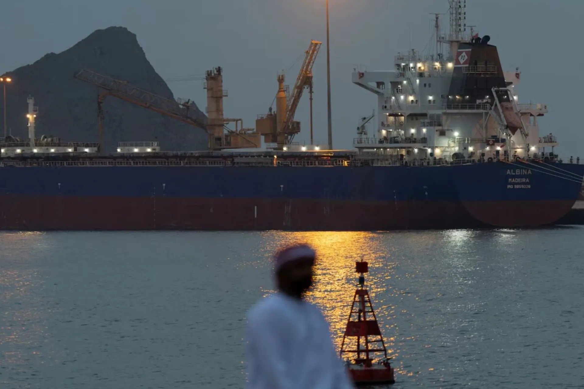  «یک مرد در کنار ساحل نشسته است، در حالی که یک کشتی در داخل بندر سلطان قابوس در لنگرگاه قرار دارد، در میانه درگیری ایالات متحده و اسرائیل با ایران