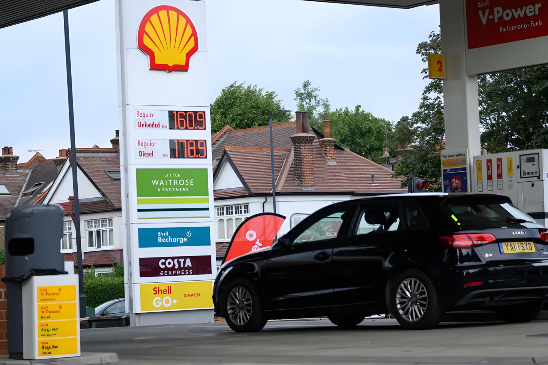 تابلوی قیمت سوخت در پمپ بنزین شل (Shell) در کنار خودروی سیاه