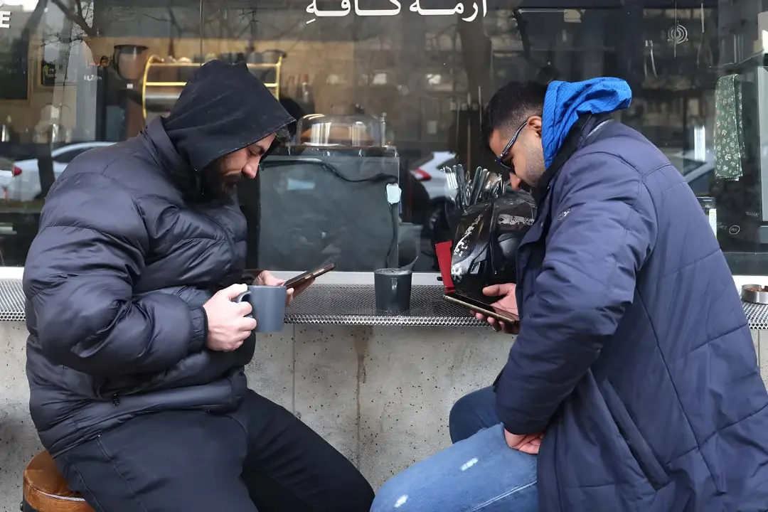 دو مرد با لباس‌های گرم در حال استفاده از گوشی‌های هوشمند و اینترنت در کافه ای در ایران